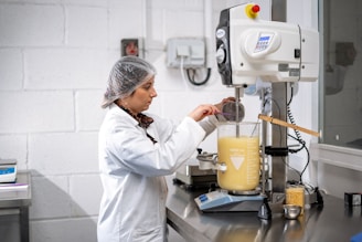 A person dressed in a lab coat and hairnet is working in a laboratory environment. They are using a large beaker filled with a yellow liquid, placed on an electronic balance. The individual is adding an ingredient using a small cup while monitoring the process.