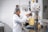 A person dressed in a lab coat and hairnet is working in a laboratory environment. They are using a large beaker filled with a yellow liquid, placed on an electronic balance. The individual is adding an ingredient using a small cup while monitoring the process.