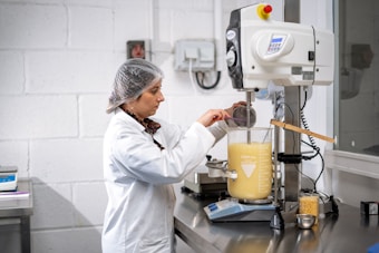 A person dressed in a lab coat and hairnet is working in a laboratory environment. They are using a large beaker filled with a yellow liquid, placed on an electronic balance. The individual is adding an ingredient using a small cup while monitoring the process.