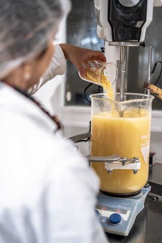 A person wearing a lab coat and hairnet is pouring yellow granules from a glass container into a large beaker filled with a yellow liquid. The setup is on a digital scale, with a blending or mixing machine connected to the beaker.