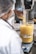 A person wearing a lab coat and hairnet is pouring yellow granules from a glass container into a large beaker filled with a yellow liquid. The setup is on a digital scale, with a blending or mixing machine connected to the beaker.