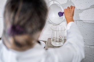A person wearing a lab coat is pouring a white liquid from a container with a purple handle into a transparent glass beaker. The beaker is placed on a metal stand against a textured white wall.