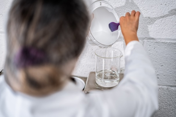 A person wearing a lab coat is pouring a white liquid from a container with a purple handle into a transparent glass beaker. The beaker is placed on a metal stand against a textured white wall.