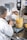 A person wearing a lab coat and hairnet is working with a large industrial mixer in a laboratory setting. They are pouring a substance, possibly corn kernels, into a transparent container filled with a creamy yellow liquid. The equipment is marked with volume measurements and is positioned on a metallic table alongside digital measurement devices.