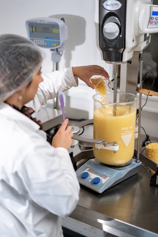 A person wearing a lab coat and hairnet is working with a large industrial mixer in a laboratory setting. They are pouring a substance, possibly corn kernels, into a transparent container filled with a creamy yellow liquid. The equipment is marked with volume measurements and is positioned on a metallic table alongside digital measurement devices.
