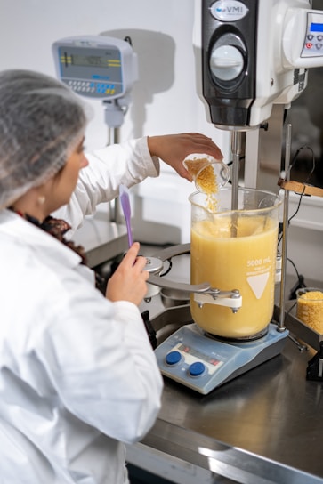 A person wearing a lab coat and hairnet is working with a large industrial mixer in a laboratory setting. They are pouring a substance, possibly corn kernels, into a transparent container filled with a creamy yellow liquid. The equipment is marked with volume measurements and is positioned on a metallic table alongside digital measurement devices.