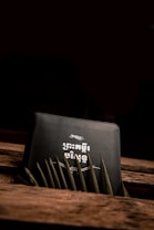 A book with an intricate design and text in an unknown script on the cover. The book is placed diagonally against a wooden surface with green leaves partially covering the bottom. The lighting is dim, creating a moody and mysterious atmosphere.