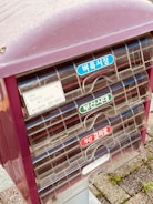 A close-up view of a set of mailboxes with colorful labels in Korean. The mailboxes are housed in a purple casing, and the labels are blue, green, and red. The ground is paved with textured stones, and some small patches of grass are visible.