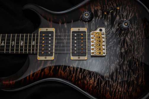 A detailed close-up of an electric guitar showcases the body and strings. The guitar features two humbucker pickups with golden screws. The bridge is metallic with several strings attached, and the fretboard shows intricate inlay designs. The guitar's body has a dark, glossy finish with a unique wood grain pattern.