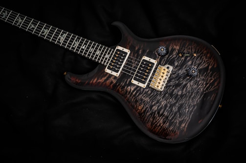 A sleek electric guitar with a richly patterned dark wood finish and detailed inlay work on the fretboard. The guitar's hardware includes dual humbucker pickups and a gold bridge, with control knobs for adjusting sound settings. It rests on a dark fabric background, enhancing the contrast and highlighting the instrument's design.