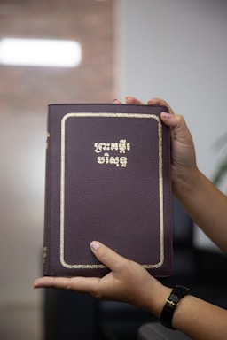 A person is holding a brown book with gold lettering on the cover. The hands have manicured nails and a watch is visible on one wrist. The background is blurred, suggesting an indoor setting.