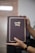 A person is holding a brown book with gold lettering on the cover. The hands have manicured nails and a watch is visible on one wrist. The background is blurred, suggesting an indoor setting.