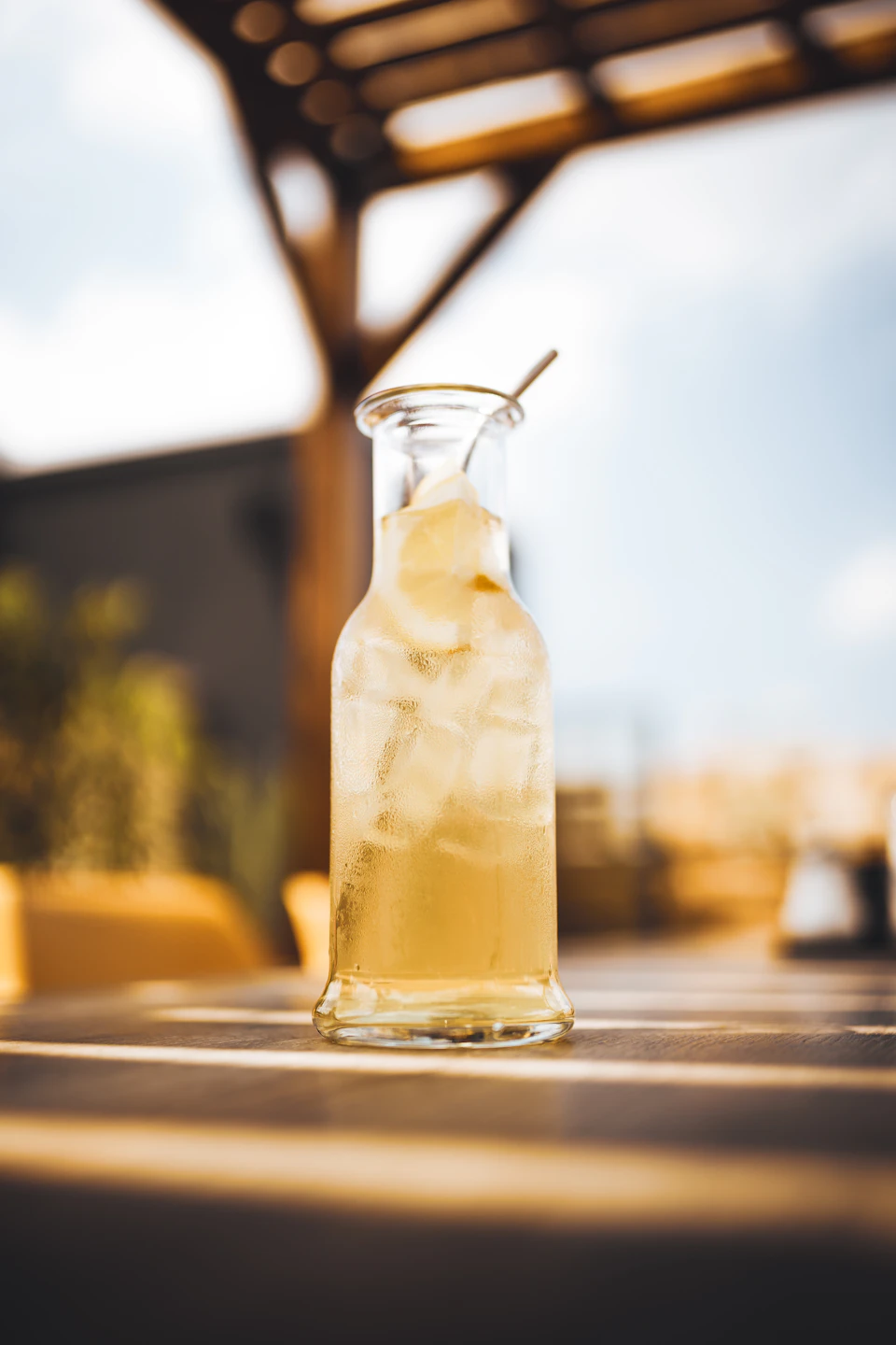 Tall glass pitcher filled with ice cubes and yellow liquid on a wooden outdoor table