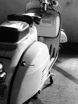 A vintage scooter is parked indoors, showcasing a classic design with a saddle seat and chrome details. A sticker on the glove box reads 'Loud Pipes Save Lives'. The lighting highlights the curves and details of the scooter, casting shadows on the concrete floor.