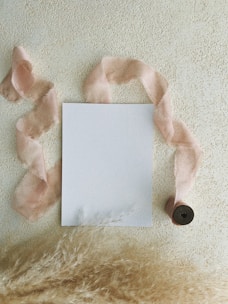 A blank piece of paper is centered on a textured, beige background. A soft, pink ribbon meanders around the paper extending from a spool at the bottom right corner. Wispy pampas grass is positioned along the bottom edge, enhancing the composition with a natural, delicate touch.