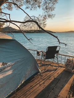 A scenic lakeside setting at sunset with a camping tent pitched on wooden decking. A single empty chair faces the tranquil water, surrounded by trees. The warm glow of the sunset casts a peaceful atmosphere over the calm lake.
