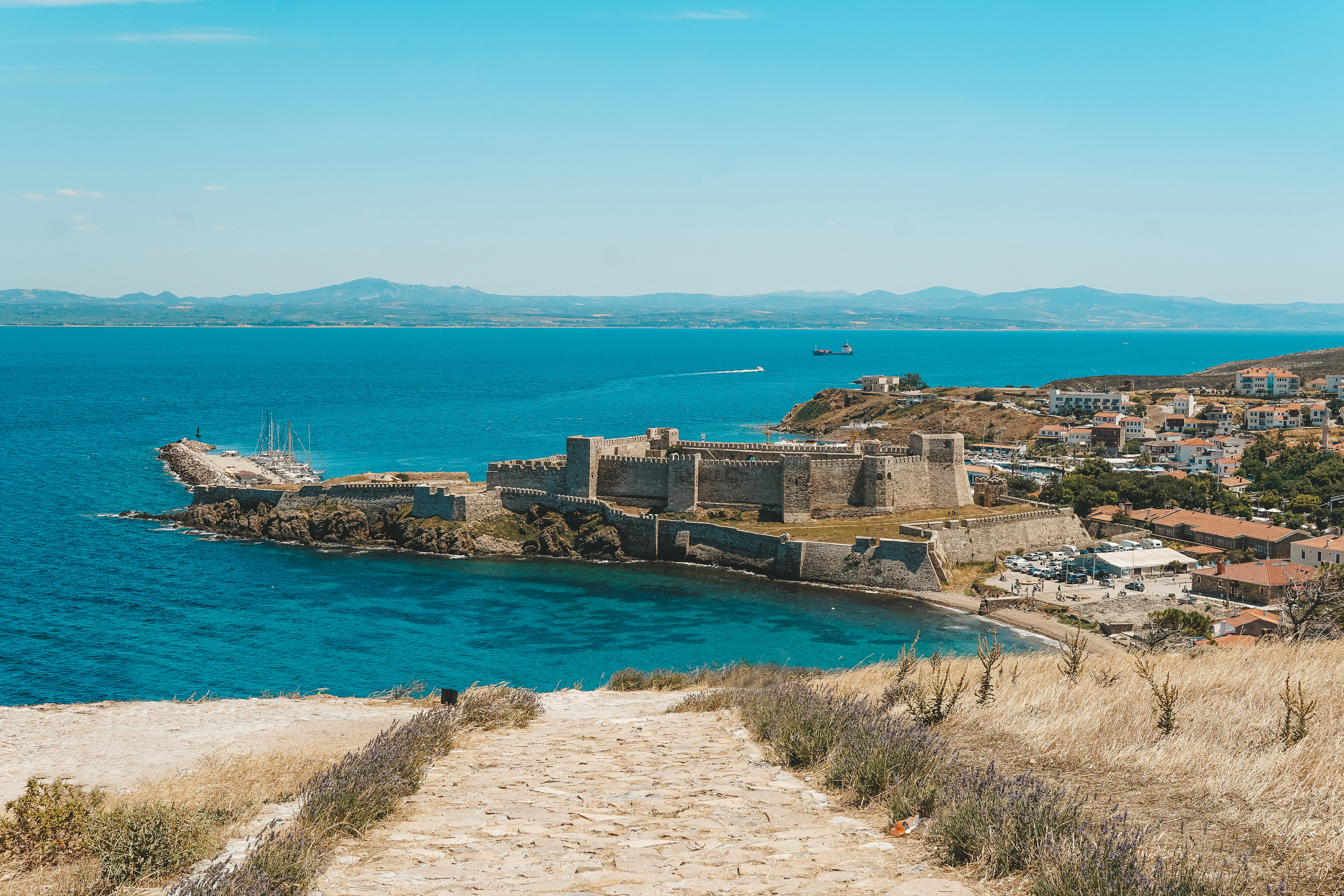 Bozcaada's fortress by the harbour, a red-roofed town and blue sea, with the mainland hills beyond
