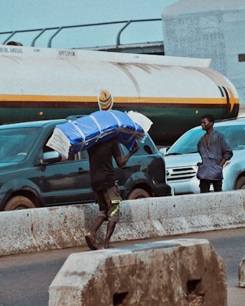 A person carrying a large, wrapped blue parcel on their head walks along a bustling roadway. Several vehicles, including a dark blue van and a white SUV, are visible. A concrete barrier separates the pedestrian from the traffic. An onlooker stands nearby, observing.