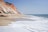 A pristine beach landscape features soft sandy shores meeting clear ocean waves. Tall, dramatic cliffs with warm reddish and white hues rise majestically along the coastline. The sky is clear, suggesting a calm, sunny day.