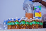 A person is pouring liquid, possibly juice or water, into a large glass pitcher. Various bottled drinks are neatly arranged on a table, including beverages with green labels and some water bottles with blue caps. A carton of multi-fruit nectar with vibrant fruit illustrations is visible, suggesting a setting for serving drinks.