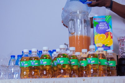 A person is pouring liquid, possibly juice or water, into a large glass pitcher. Various bottled drinks are neatly arranged on a table, including beverages with green labels and some water bottles with blue caps. A carton of multi-fruit nectar with vibrant fruit illustrations is visible, suggesting a setting for serving drinks.