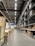 A large warehouse with high ceilings and rows of tall metal shelves filled with cardboard boxes. The space is well-lit with fluorescent lights, and there are signs and partitions on the left side, suggesting an organized industrial or retail environment.