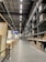 A large warehouse with high ceilings and rows of tall metal shelves filled with cardboard boxes. The space is well-lit with fluorescent lights, and there are signs and partitions on the left side, suggesting an organized industrial or retail environment.