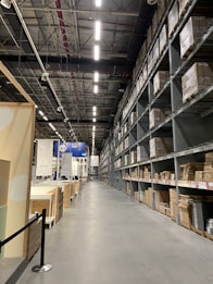 A large warehouse with high ceilings and rows of tall metal shelves filled with cardboard boxes. The space is well-lit with fluorescent lights, and there are signs and partitions on the left side, suggesting an organized industrial or retail environment.