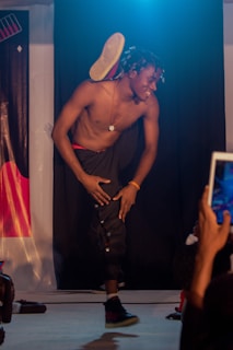 A male dancer energetically performing on stage, balancing one leg against his shoulder. He is shirtless, wearing dark pants, and surrounded by an audience capturing the moment. The stage lighting casts a dramatic ambiance, highlighting his movement.