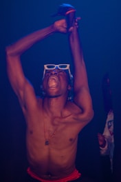 A person is expressing intense emotion with their mouth open wide, wearing white-framed sunglasses, and raising their arms above their head. The scene is set in low lighting with a blue hue. They are shirtless, with a red waistband, and wearing a necklace with a pendant.