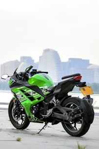 A sleek, green and black sports motorcycle is parked on a paved surface near a waterfront. In the background, there are modern high-rise buildings with various architectural designs, suggesting an urban setting. The motorcycle's design is aerodynamic with a prominent fairing, and it features a side stand and a visible exhaust system.