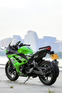 A sleek, green and black sports motorcycle is parked on a paved surface near a waterfront. In the background, there are modern high-rise buildings with various architectural designs, suggesting an urban setting. The motorcycle's design is aerodynamic with a prominent fairing, and it features a side stand and a visible exhaust system.