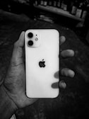 A hand holding a white smartphone with a prominent logo featuring an apple with a bite taken out of it. The background appears to be a blurred indoor setting with shelves lined with bottles.