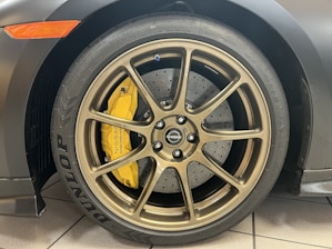 A close-up view of a car wheel featuring a bronze-colored alloy rim with a visible Nissan logo at the center. The tire has Dunlop and other branding inscribed, and behind the rim there is a yellow brake caliper. The brake disc appears drilled and ventilated.