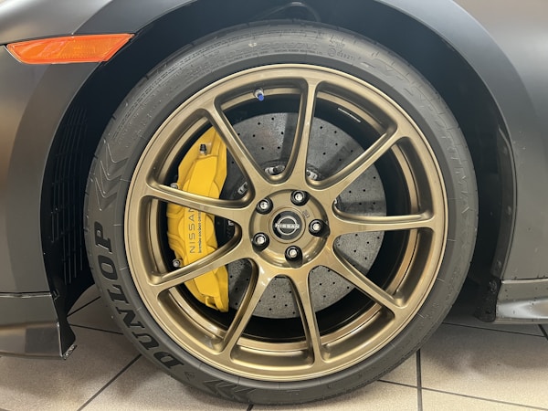 A close-up view of a car wheel featuring a bronze-colored alloy rim with a visible Nissan logo at the center. The tire has Dunlop and other branding inscribed, and behind the rim there is a yellow brake caliper. The brake disc appears drilled and ventilated.