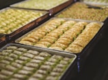 Several trays filled with pieces of baklava, a traditional dessert made with layers of filo pastry, nuts, and syrup or honey. The baklava is neatly arranged, with a golden brown color and sprinkled with crushed pistachios.