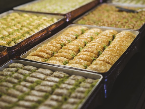 Several trays filled with pieces of baklava, a traditional dessert made with layers of filo pastry, nuts, and syrup or honey. The baklava is neatly arranged, with a golden brown color and sprinkled with crushed pistachios.