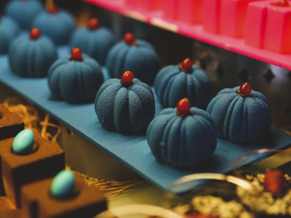 A display of intricately designed dark blue desserts with a velvety texture, topped with a small red garnish, set on a matching blue tray. In the foreground, a hint of chocolate squares adorned with bright teal ovals is visible.