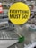A yellow circular sign with black text saying 'EVERYTHING MUST GO!' is prominently displayed in the center. In the background, there is an indoor retail store setting with shelves, a staircase, and shopping carts. Several other sale signs are visible in the distance, indicating that the store is likely closing down.