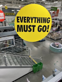 A yellow circular sign with black text saying 'EVERYTHING MUST GO!' is prominently displayed in the center. In the background, there is an indoor retail store setting with shelves, a staircase, and shopping carts. Several other sale signs are visible in the distance, indicating that the store is likely closing down.