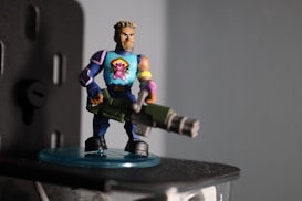 A toy figurine resembling a muscular character holding a large, green weapon stands on a small transparent base. The character has short hair, a blue shirt with a cartoon emblem featuring a pink creature, and dark blue pants. The figurine appears detailed with additional vibrant colors on its armband and shoes.