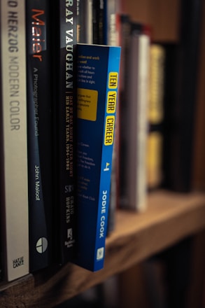 A close-up view of a shelf lined with several books. One book has a bright blue spine with the title 'Ten Year Career' by Jodie Cook written in yellow block letters. Surrounding it are various other books with titles visible, including 'Gray Vaughan' and 'Maier'. The books are neatly arranged, creating a sense of order.