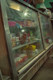 A glass display case containing various packaged food items, such as beverages, snacks, and grocery goods. The case appears slightly foggy or aged, giving it an old-fashioned look. Bags of chips, packets of cookies, and cartons of juice are visible on the shelves. Above the display case, suspended plastic bags with additional food items can be seen.
