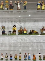 A display of small ceramic figurines arranged on shelves, depicting various characters in traditional attire. The figures are in rows on transparent shelves against a textured wall, featuring a range of vibrant colors and intricate details in their costumes and poses.