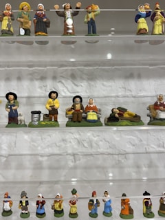 A display of small ceramic figurines arranged on shelves, depicting various characters in traditional attire. The figures are in rows on transparent shelves against a textured wall, featuring a range of vibrant colors and intricate details in their costumes and poses.