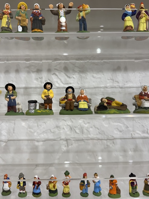 A display of small ceramic figurines arranged on shelves, depicting various characters in traditional attire. The figures are in rows on transparent shelves against a textured wall, featuring a range of vibrant colors and intricate details in their costumes and poses.