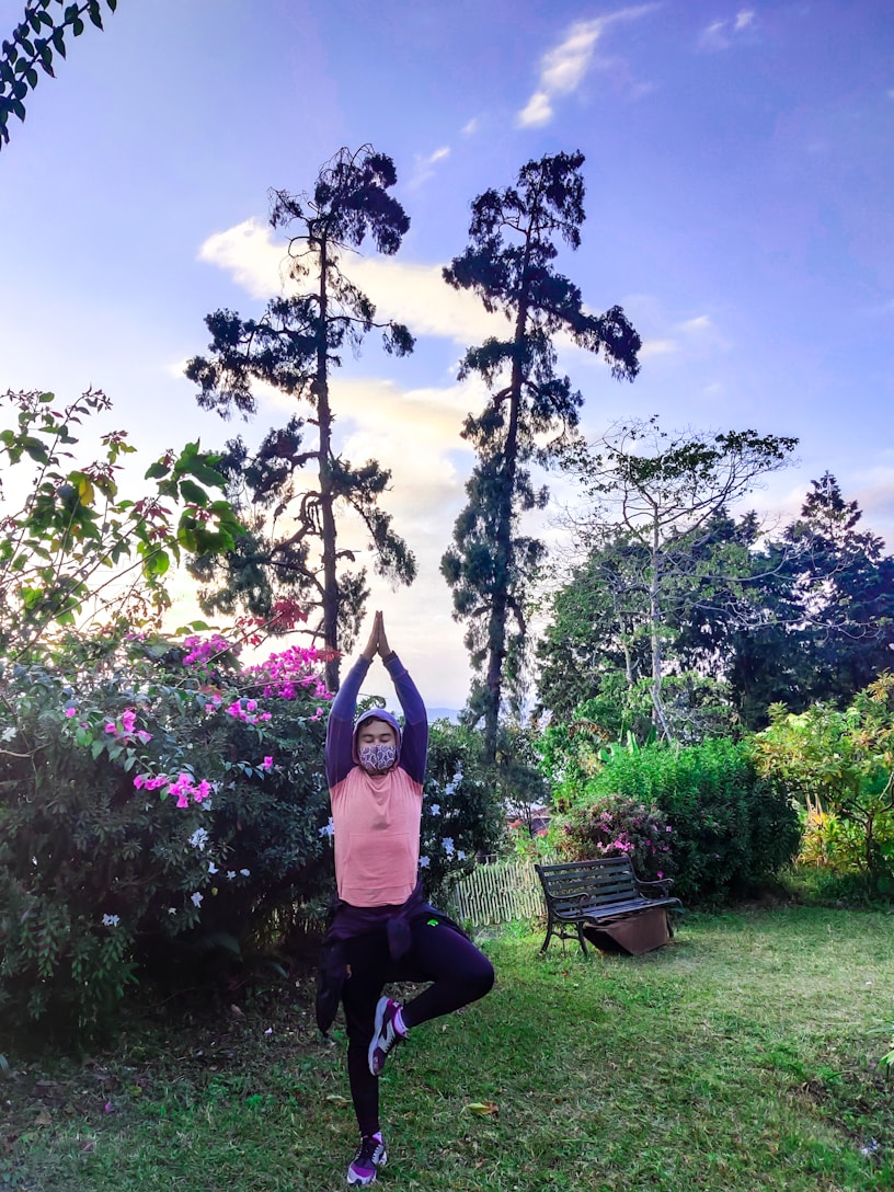 A person practices a yoga pose in a lush garden surrounded by vibrant pink flowers and tall trees under a clear sky. They are wearing a face mask and balancing on one foot while raising arms overhead in a serene outdoor setting.