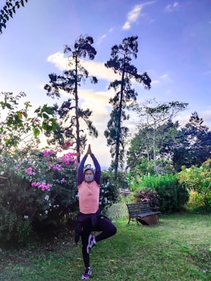 A person practices a yoga pose in a lush garden surrounded by vibrant pink flowers and tall trees under a clear sky. They are wearing a face mask and balancing on one foot while raising arms overhead in a serene outdoor setting.