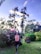 A person practices a yoga pose in a lush garden surrounded by vibrant pink flowers and tall trees under a clear sky. They are wearing a face mask and balancing on one foot while raising arms overhead in a serene outdoor setting.