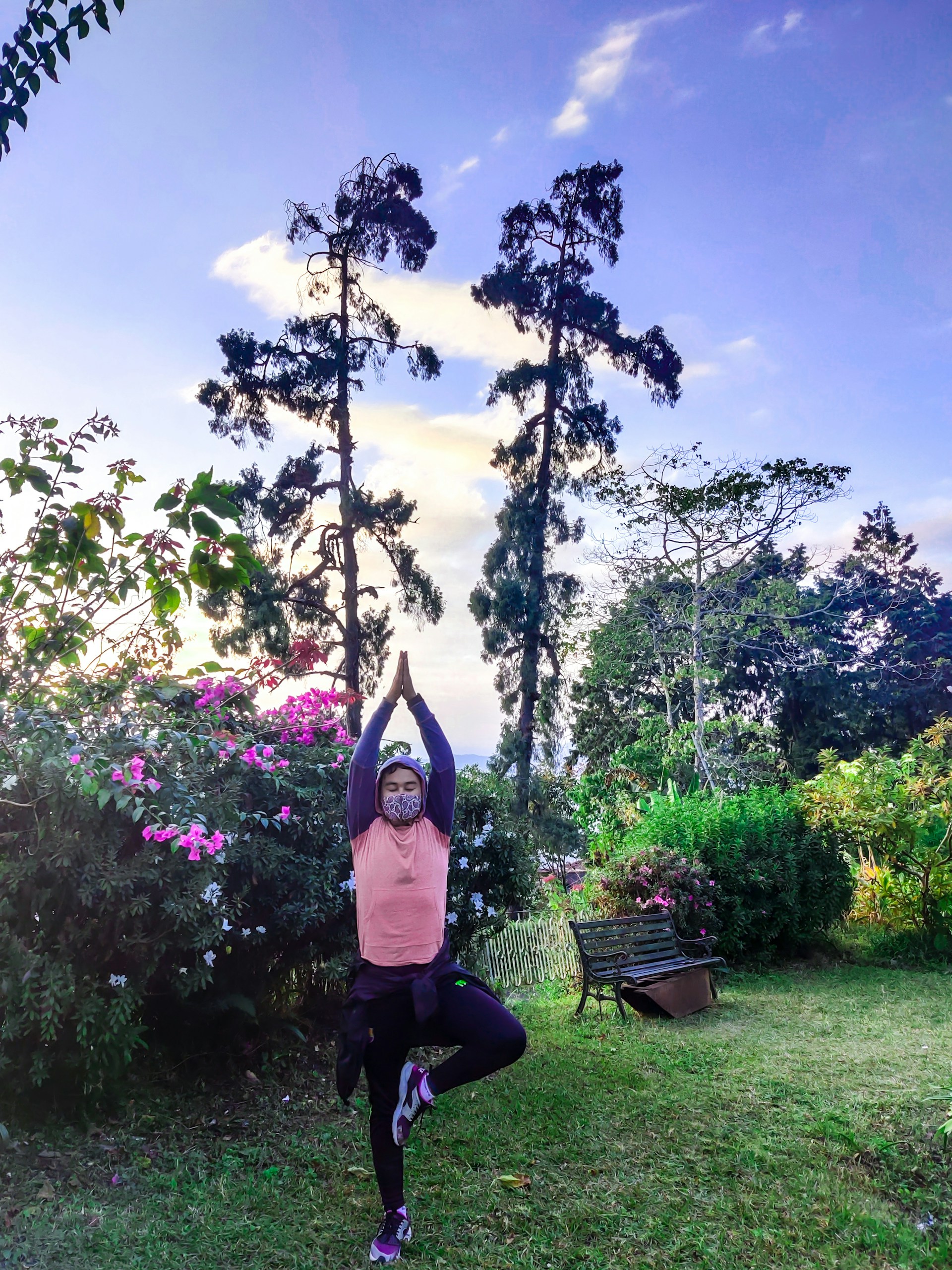 A person practices a yoga pose in a lush garden surrounded by vibrant pink flowers and tall trees under a clear sky. They are wearing a face mask and balancing on one foot while raising arms overhead in a serene outdoor setting.